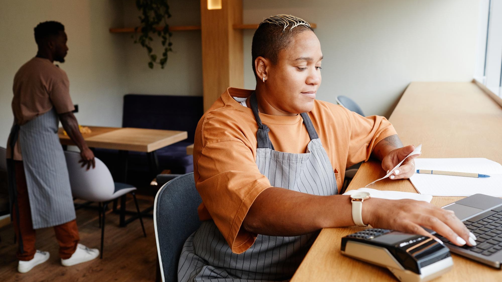 Young african american woman wearing apron sitting at table in front of laptop in modern cafe taking...