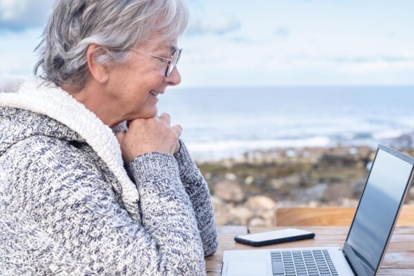 Smiling attractive mature senior adult professional business woman in remote working on laptop computer by the beach wearing eyeglasses sitting at the wooden table looking at computer screen