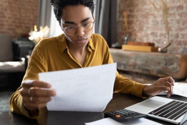 Focused young African American woman in eyeglasses looking through paper documents managing business affairs summarizing taxes planning future investments accounting alone at home office