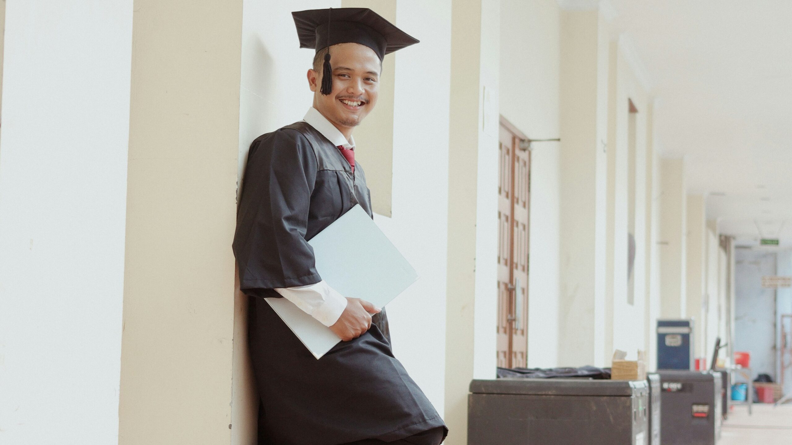 A smiling graduate in cap and gown holds a folder.