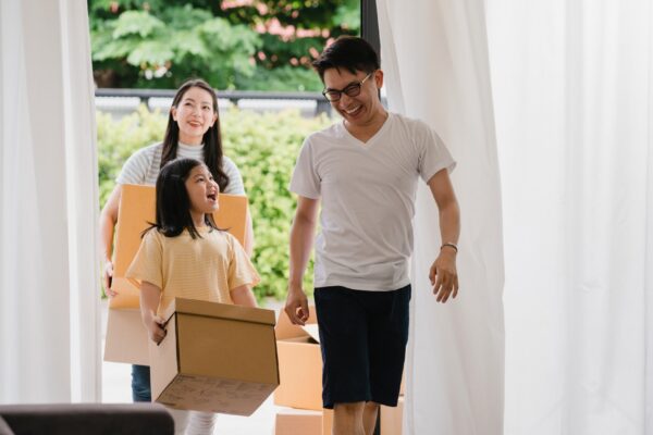 Happy Asian young family bought new house. Japanese Mom, Dad, and child smiling happy hold cardboard boxes for move object walking into big modern home. New real estate dwelling, loan and mortgage.