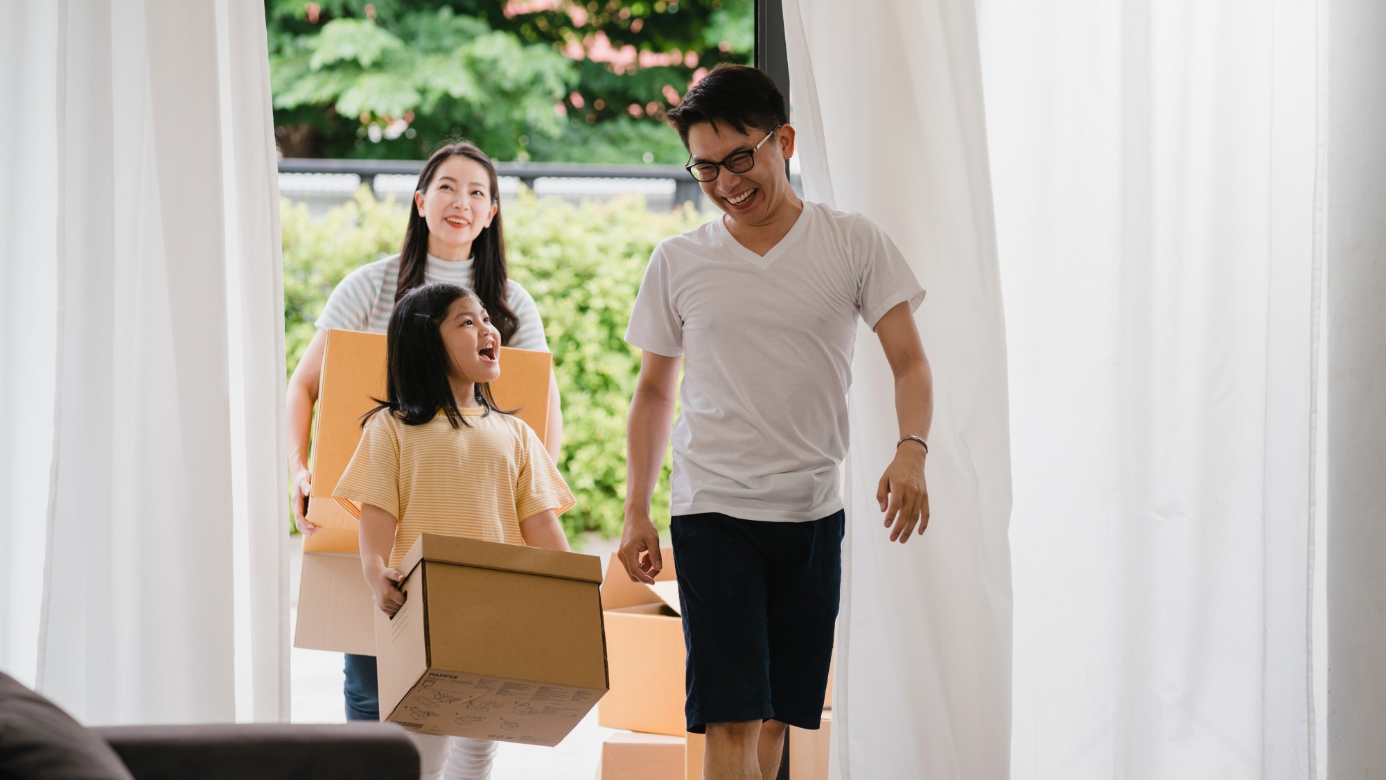 Happy Asian young family bought new house. Japanese Mom, Dad, and child smiling happy hold cardboard boxes for move object walking into big modern home. New real estate dwelling, loan and mortgage.