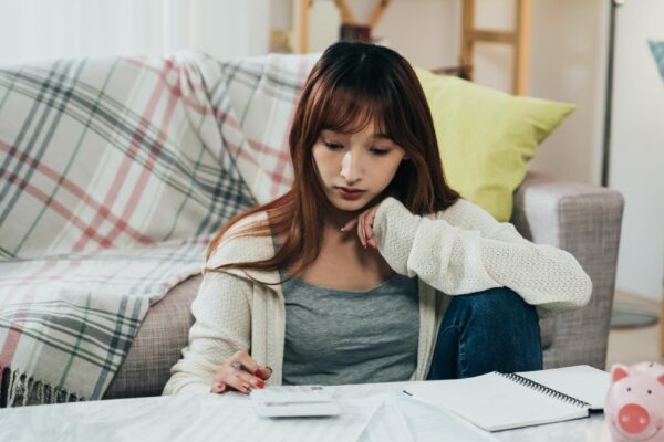 portrait concentrated asian young wife is using a calculator while thinking and planning household budget at table with a piggy bank in the living room.