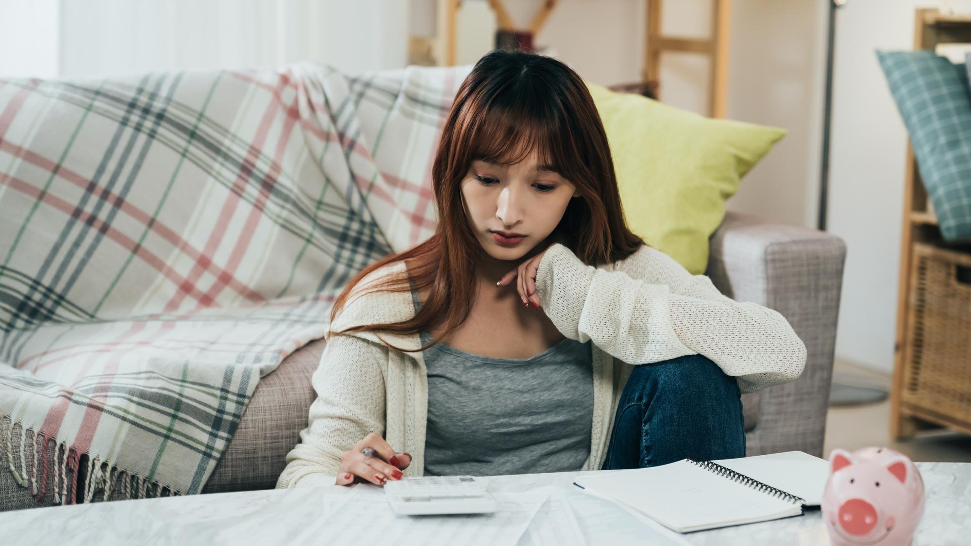 portrait concentrated asian young wife is using a calculator while thinking and planning household budget at table with a piggy bank in the living room.