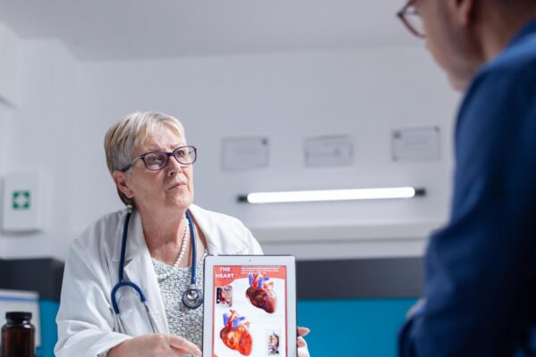 Specialist talking about cardiology diagnosis with image of heart organ on tablet to man. General practitioner showing illustration on cardiovascular system and anatomical structure to patient.
