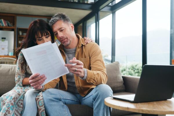Couple reading and document on couch at house for finance review budget and online payment Mature people laptop and audit paperwork on household expenses financial statement and loan application