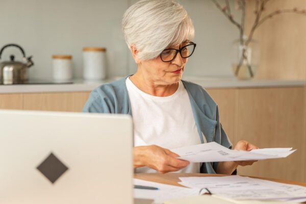 Middle aged senior woman sit with laptop and paper document pensive older mature lady reading paper