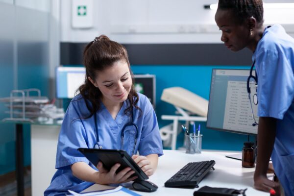 Medical team of nurses working on tablet in modern professional medical office. Smiling healthcare employee comparing data with african american coworker in hospital workplace