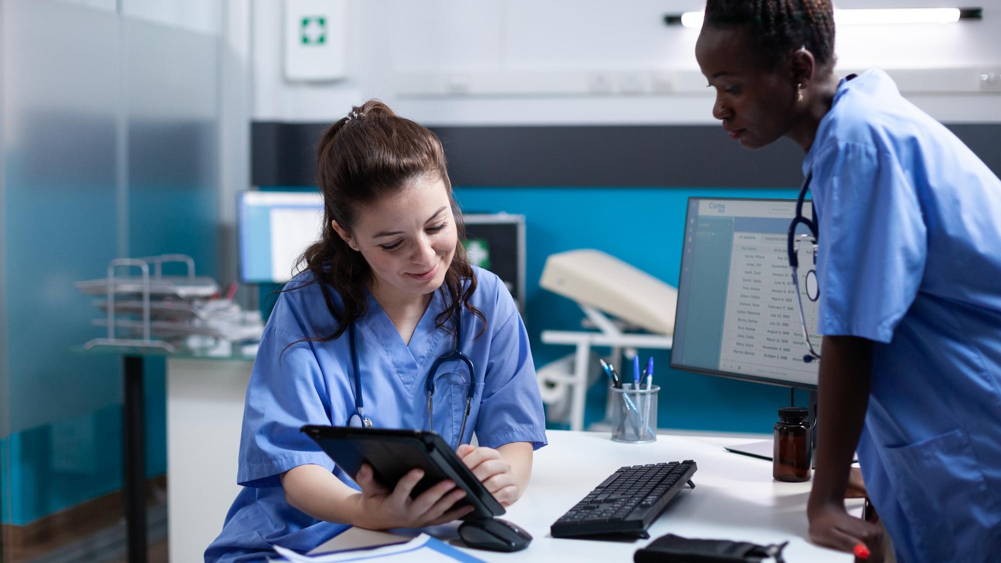 Medical team of nurses working on tablet in modern professional medical office. Smiling healthcare employee comparing data with african american coworker in hospital workplace