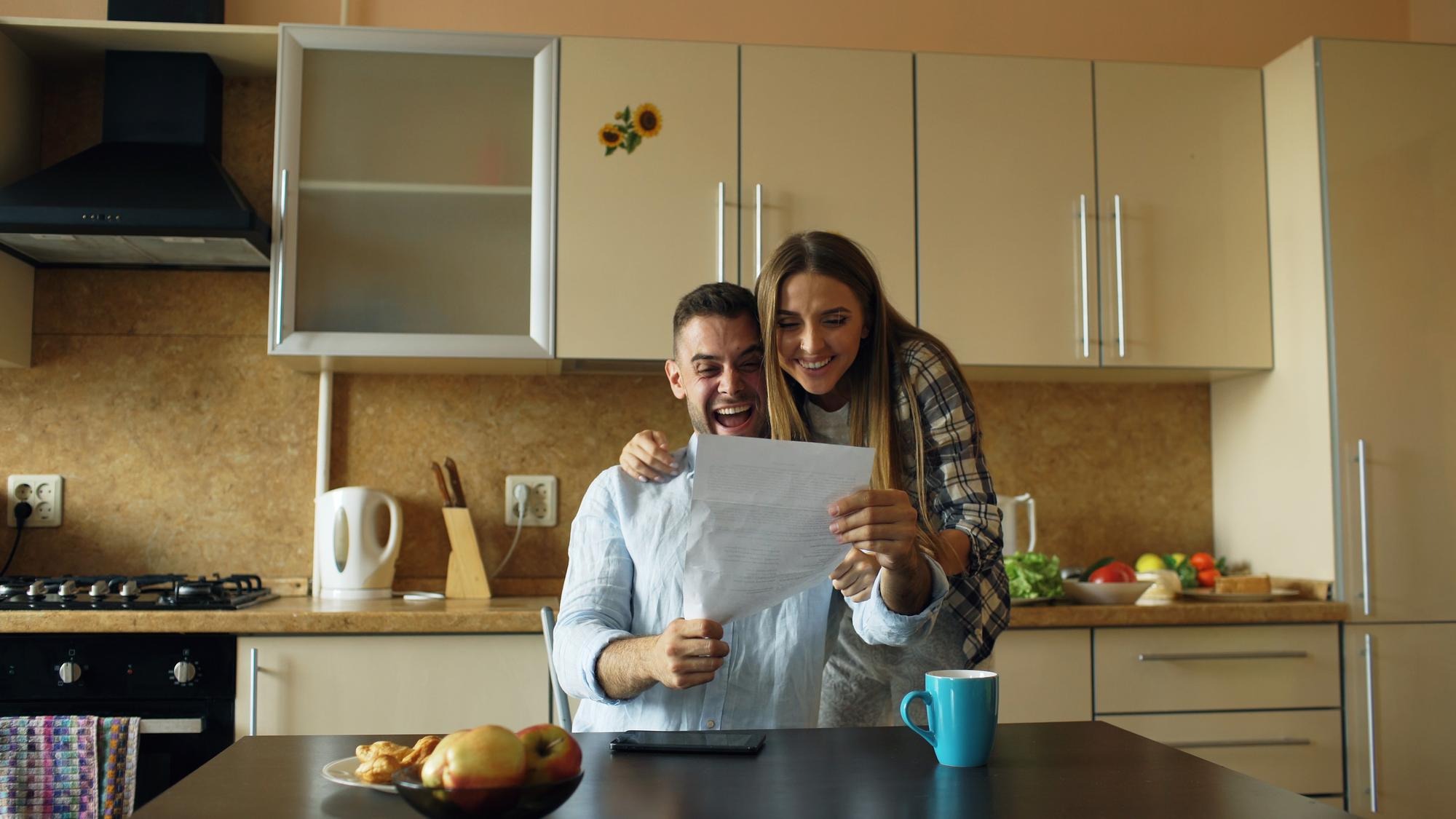 Attractive happy couple receive good news unfolding letter in the kitchen while have breakfast