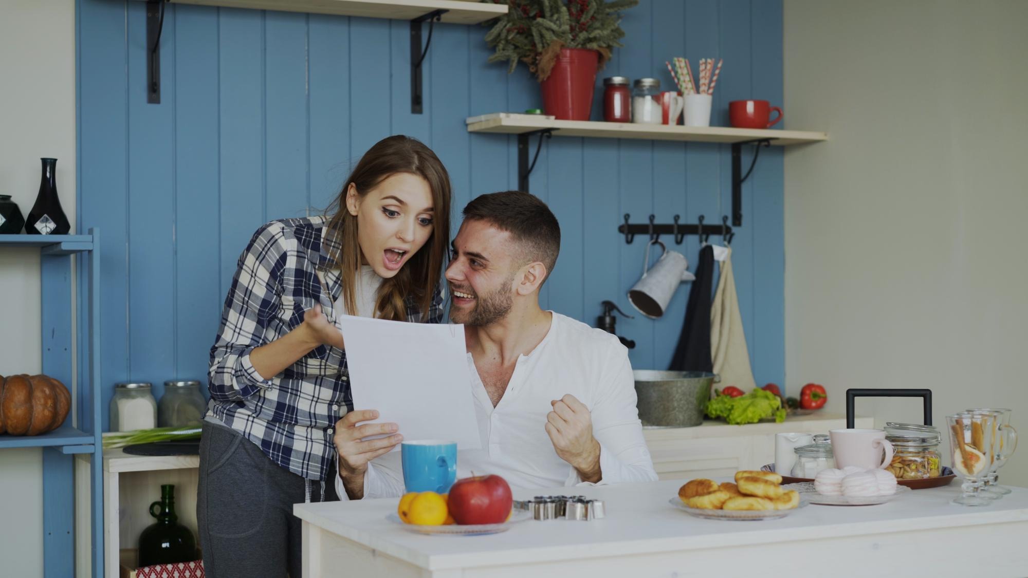 Attractive happy couple recieve good news unfolding letter in the kitchen while have breakfast