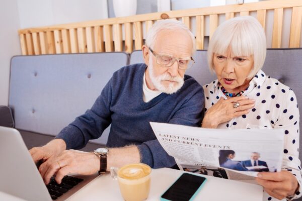 Breaking news. Amazed senior couple examining newspaper and sitting at table