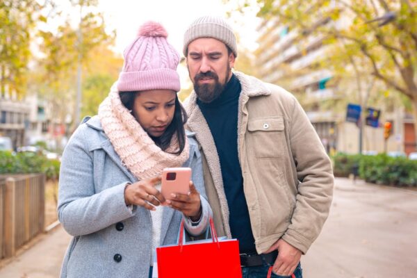 Smiling young multiracial couple holding shopping bags browsing smartphone apps