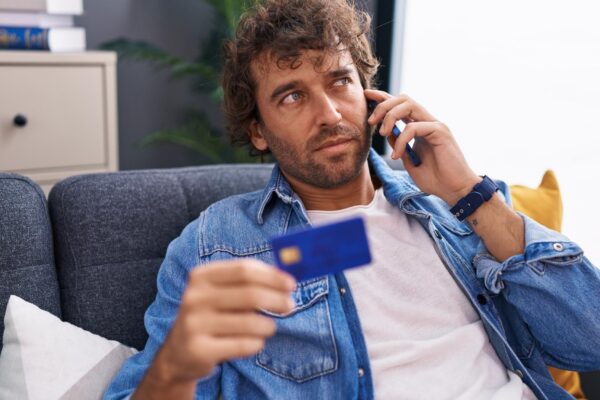 Young hispanic man talking on smartphone holding credit card sitting on sofa at home