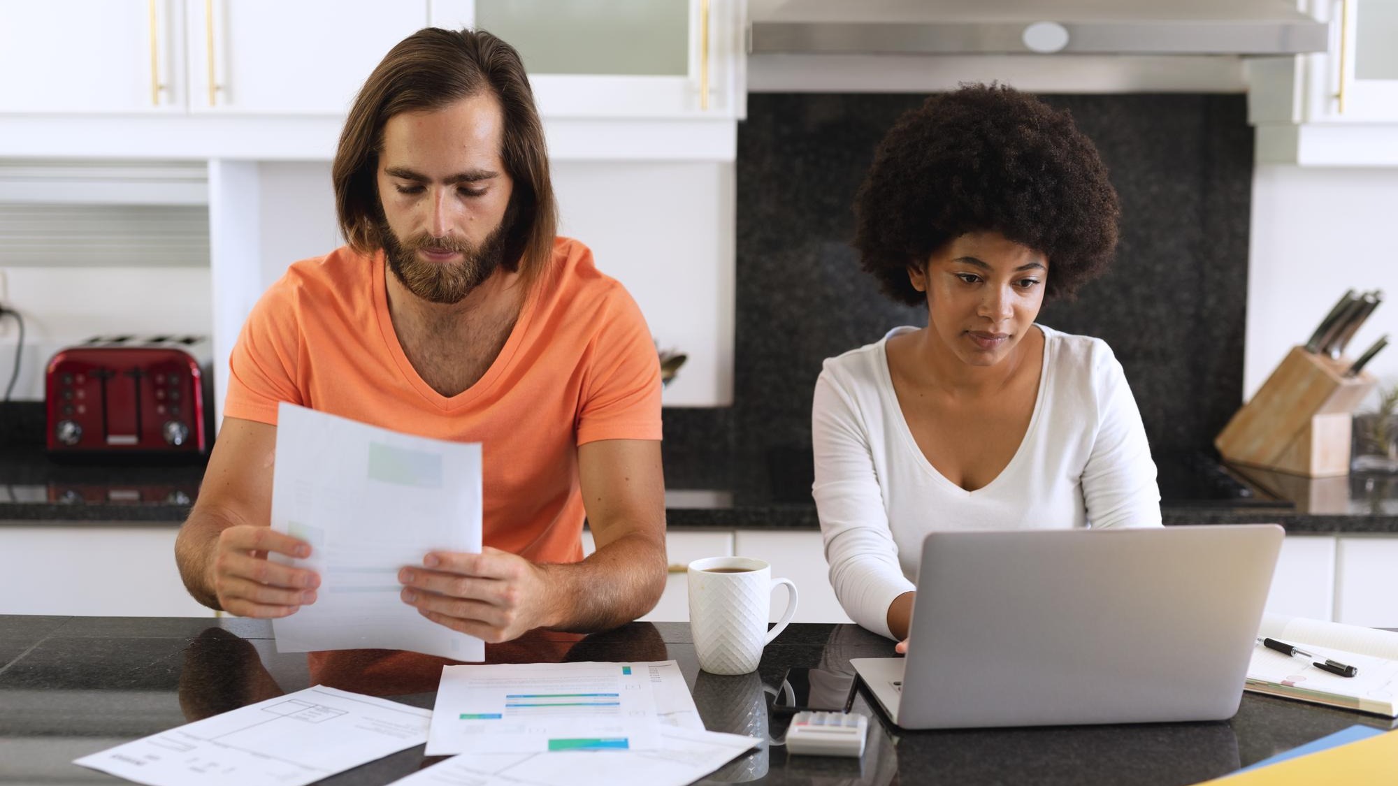 Diverse couple sitting in kitchen using laptop and paying bills. staying at home in isolation during quarantine lockdown.