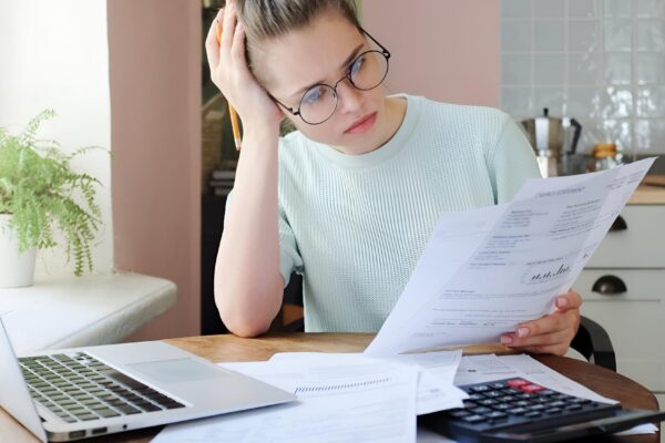 Young frustrated unhappy tired woman with financial troubles sitting at kitchen table with papers calculator and laptop computer
