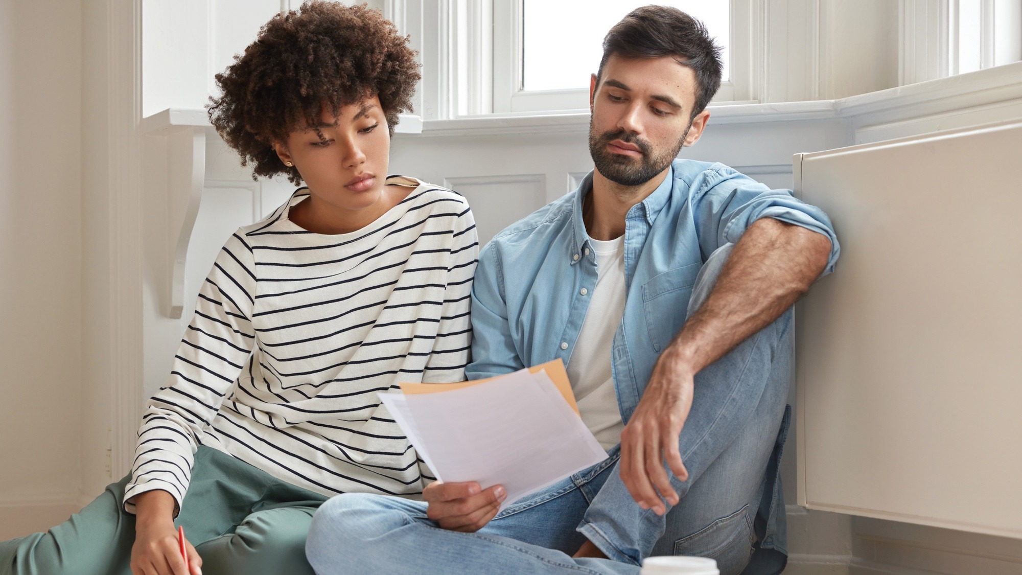 Male bearded accountant and his secretary work together in modern apartment, pose on wooden floor and discuss financial report, have serious gaze in papers, study analytics, feel comfort at home