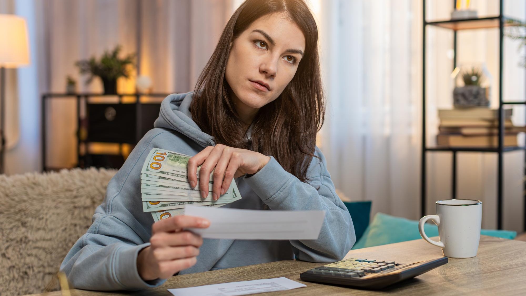 Young woman counting money while sitting on sofa with receipts and calculator on tablet at home