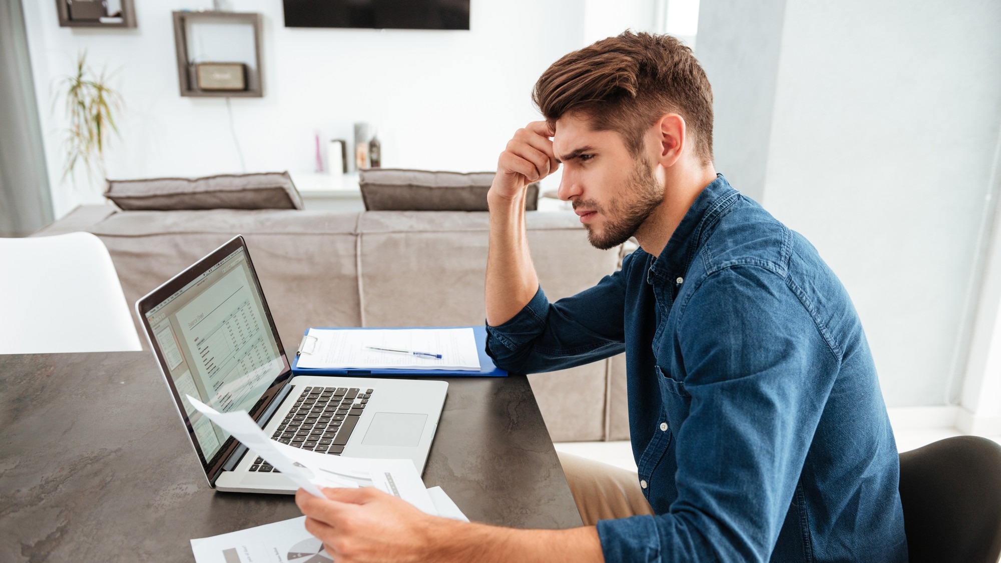 Serious concentrated man working at laptop and sitting at the table while looking at the papers and holding his head with hand