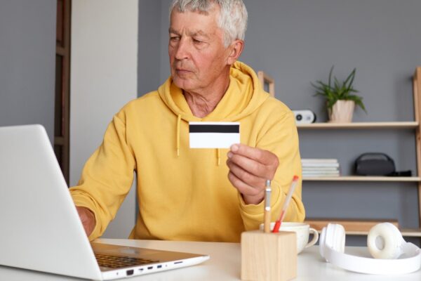 Confident modern senior man wearing yellow hoodie using laptop and credit card entering data typing on keyboard doing internet paying for purchases using internet bank