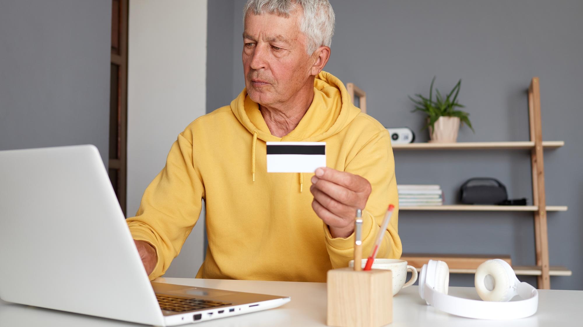 Confident modern senior man wearing yellow hoodie using laptop and credit card entering data typing on keyboard doing internet paying for purchases using internet bank