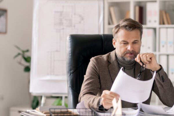 Serious or pensive mature architect looking at sketch on paper while sitting in armchair by desk in office