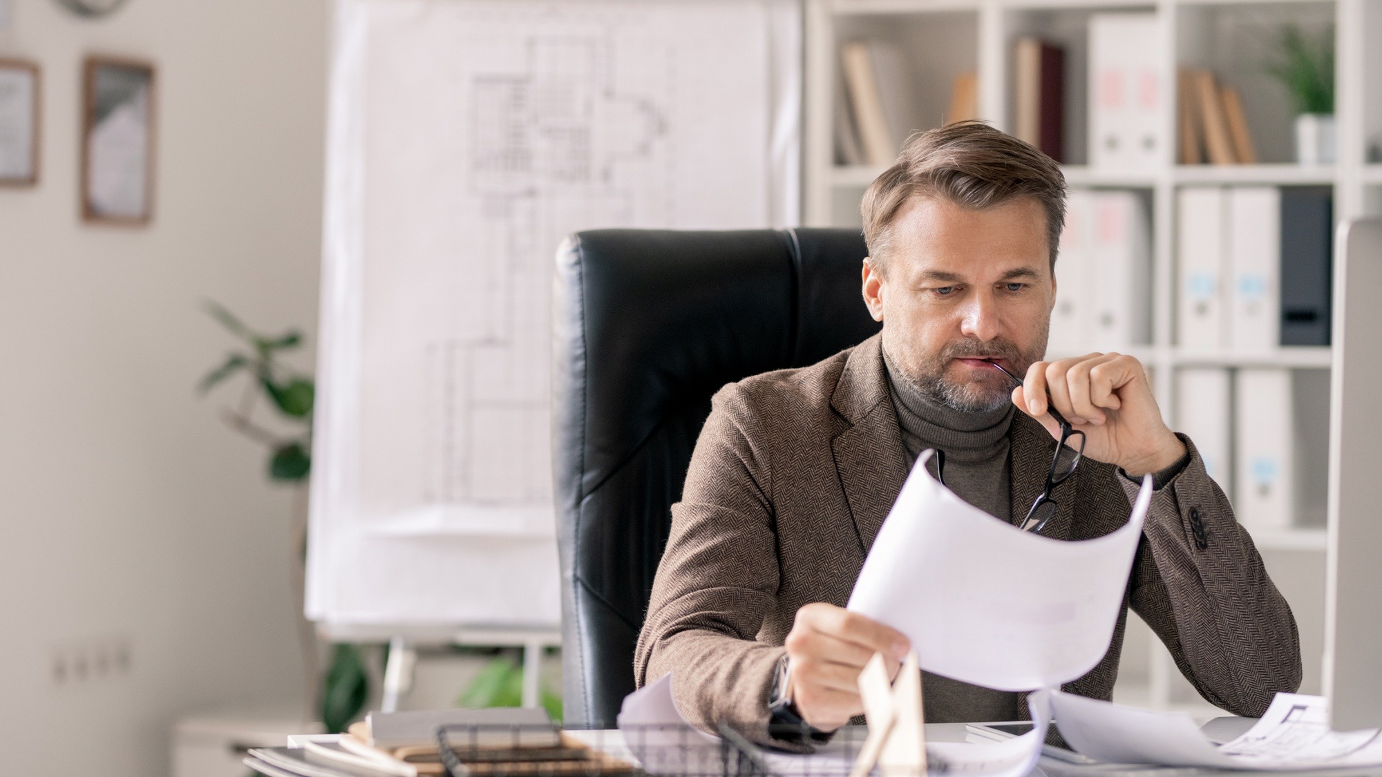 Serious or pensive mature architect looking at sketch on paper while sitting in armchair by desk in office