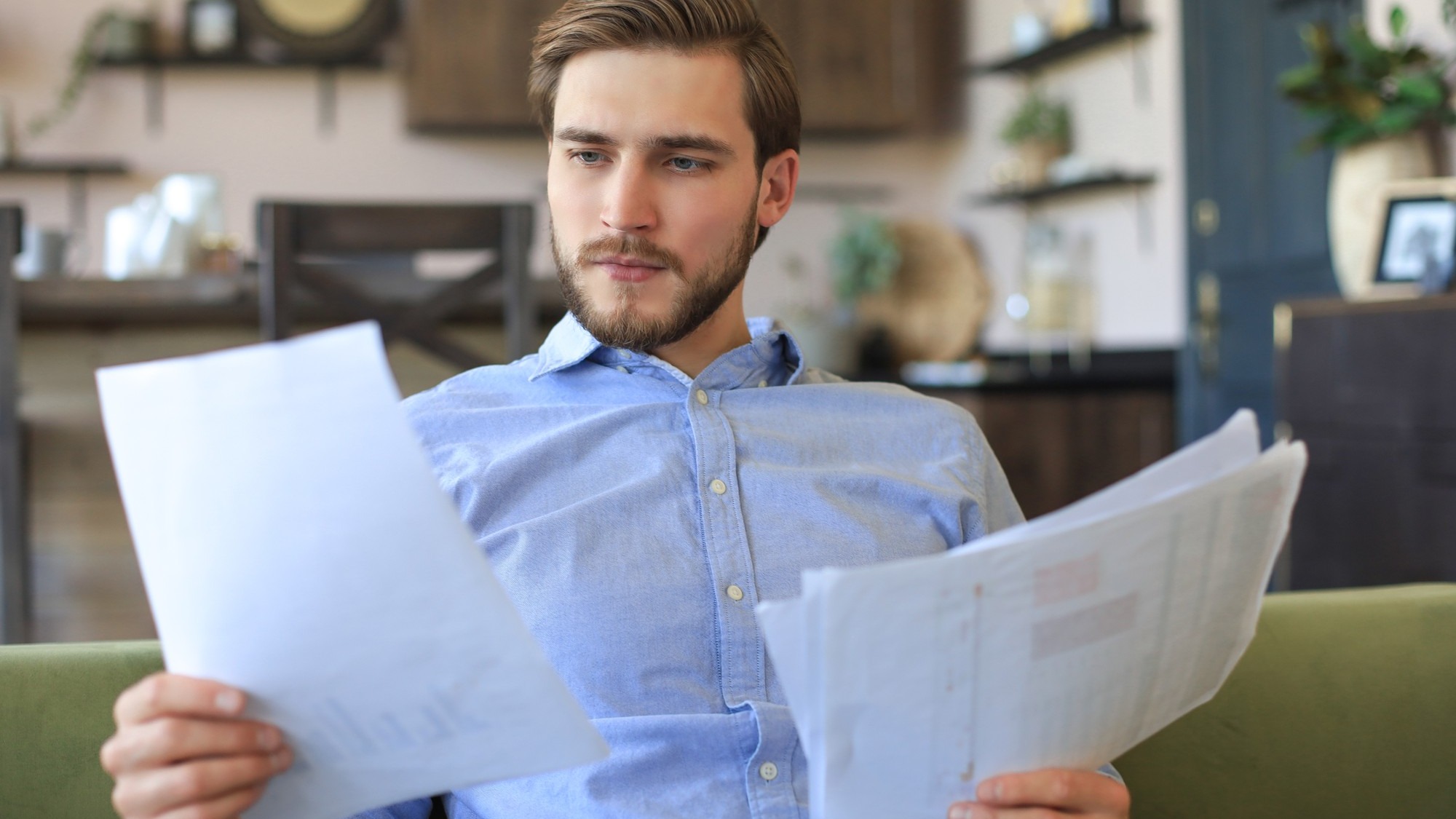 Concentrated young freelancer businessman sitting on sofa with laptop and examining documents.