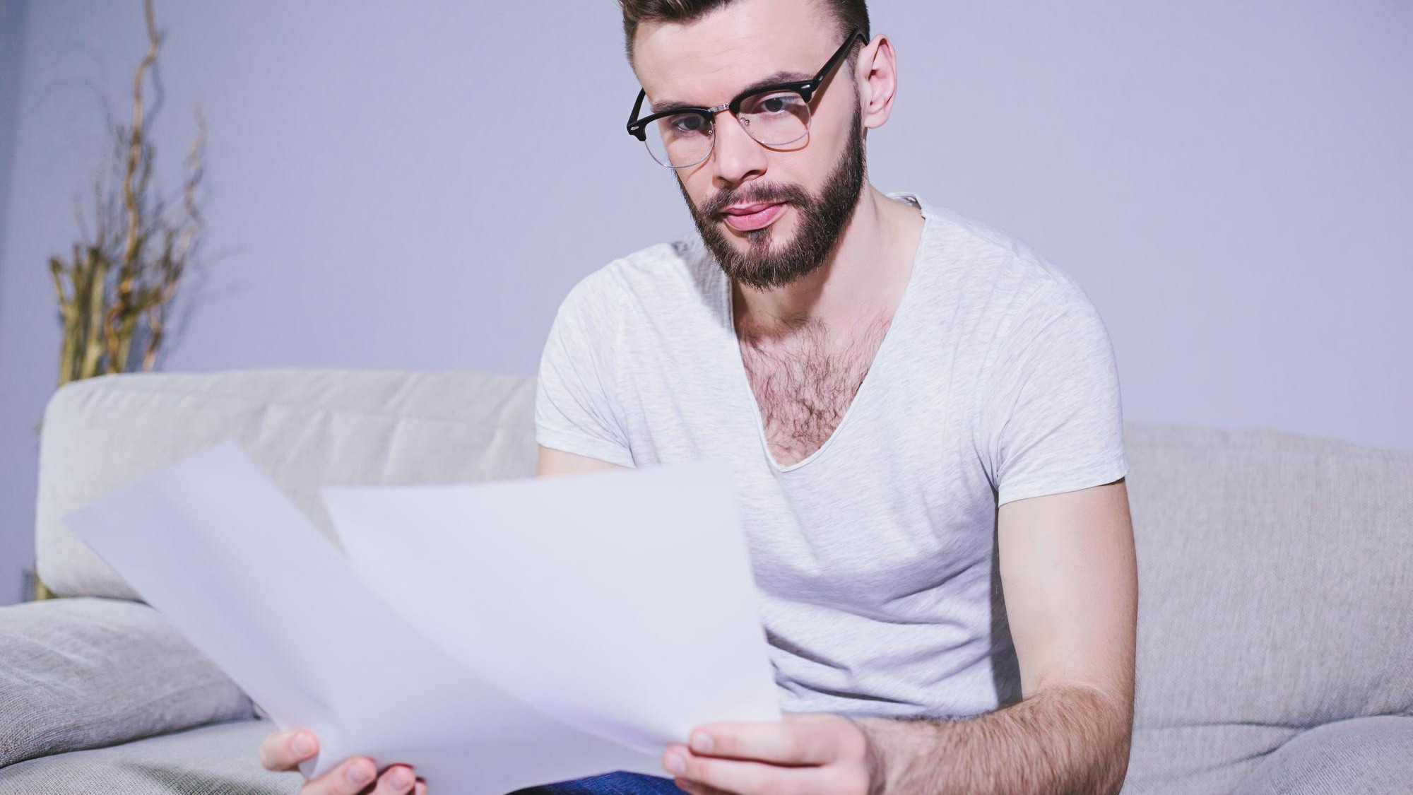 Young bearded business man with glasses working with business papers sitting at home on the couch