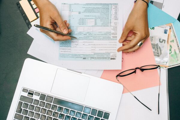 Hands writing on tax documents with laptop, glasses, and currency on desk.