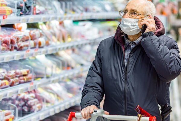 Old senior european man wearing protective facial mask pushing shopping cart and talking on the phone in the supermarket. Shopping during COVID-19 concept.