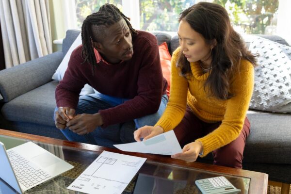 Serious diverse couple using laptop sitting on couch discussing bills and finances in living room