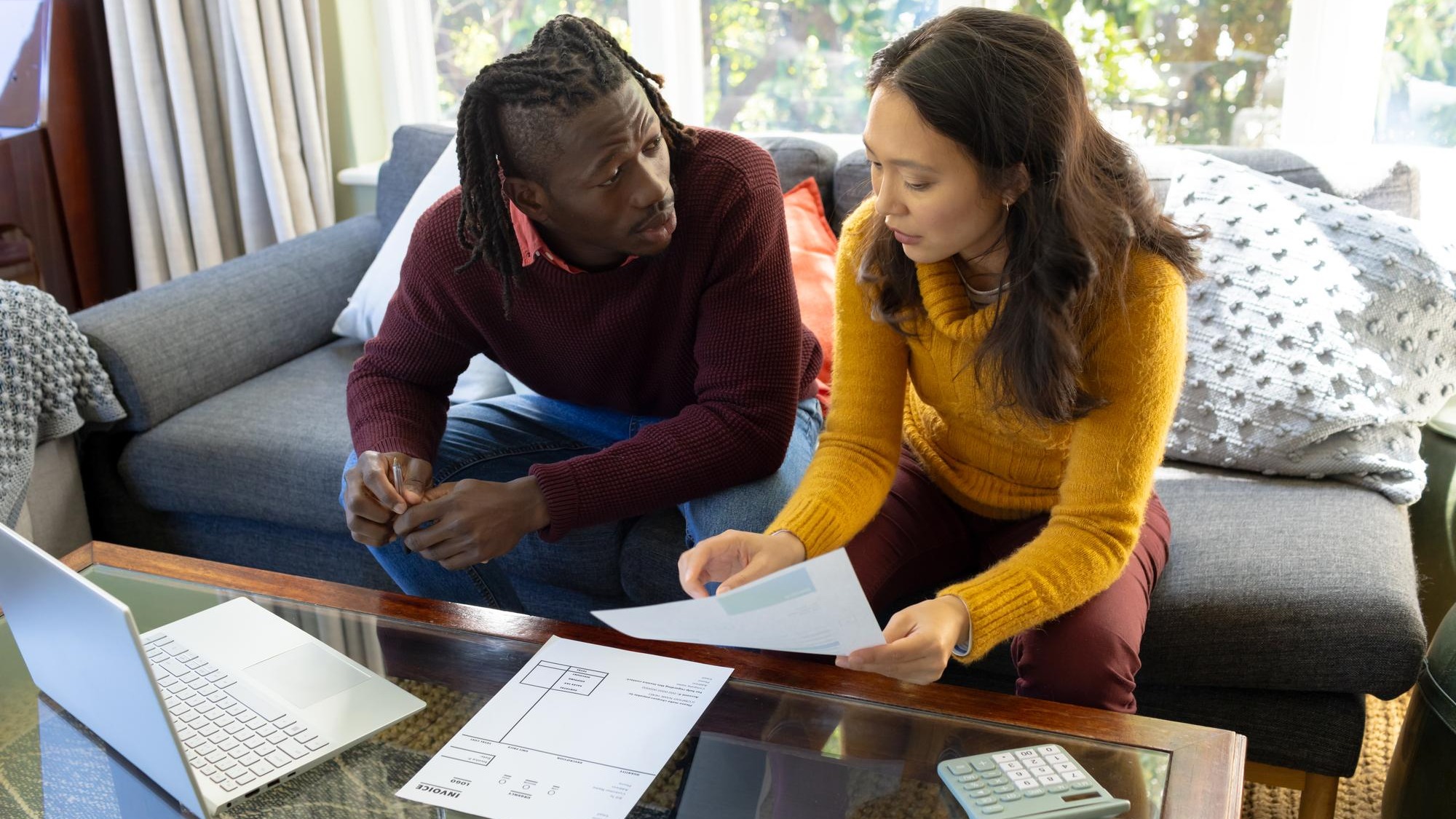Serious diverse couple using laptop sitting on couch discussing bills and finances in living room