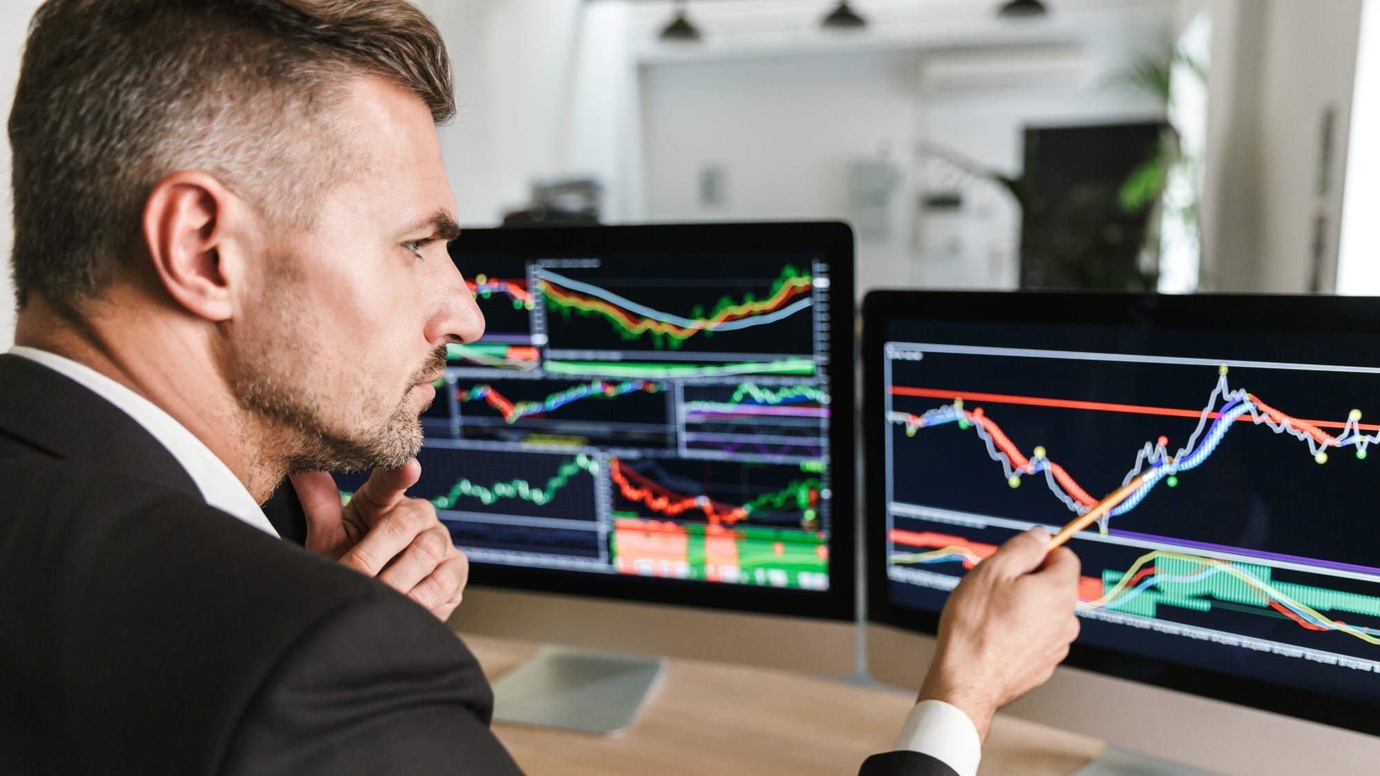 Photo of european businessman 30s wearing suit sitting at table in office and working with graphics and charts on computer