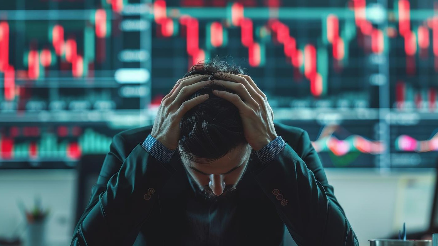 Businessperson holding head in hands at desk with falling stock charts