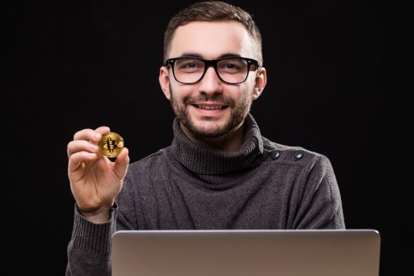 Portrait of a happy entrepreneur showing bitcoin while sitting at desk with laptop computer isolated over black