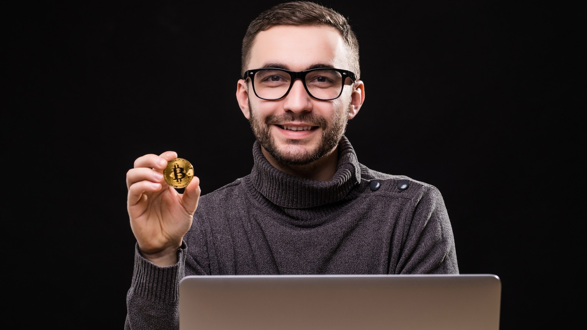 Portrait of a happy entrepreneur showing bitcoin while sitting at desk with laptop computer isolated over black