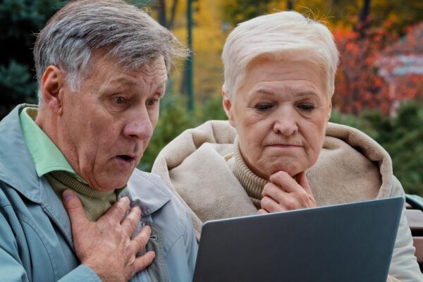 Shocked old married couple looking at laptop screen in autumn park upset elderly man and woman
