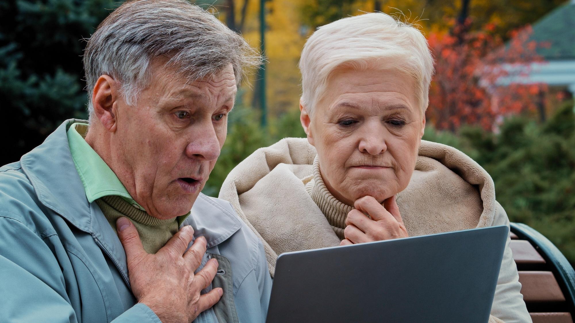 Shocked old married couple looking at laptop screen in autumn park upset elderly man and woman