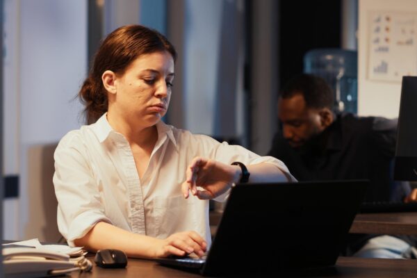 Young adult checking time on wristwatch to leave office after hours, working at night on research data to create presentation. Feeling overworked at corporate job, planning financial report.