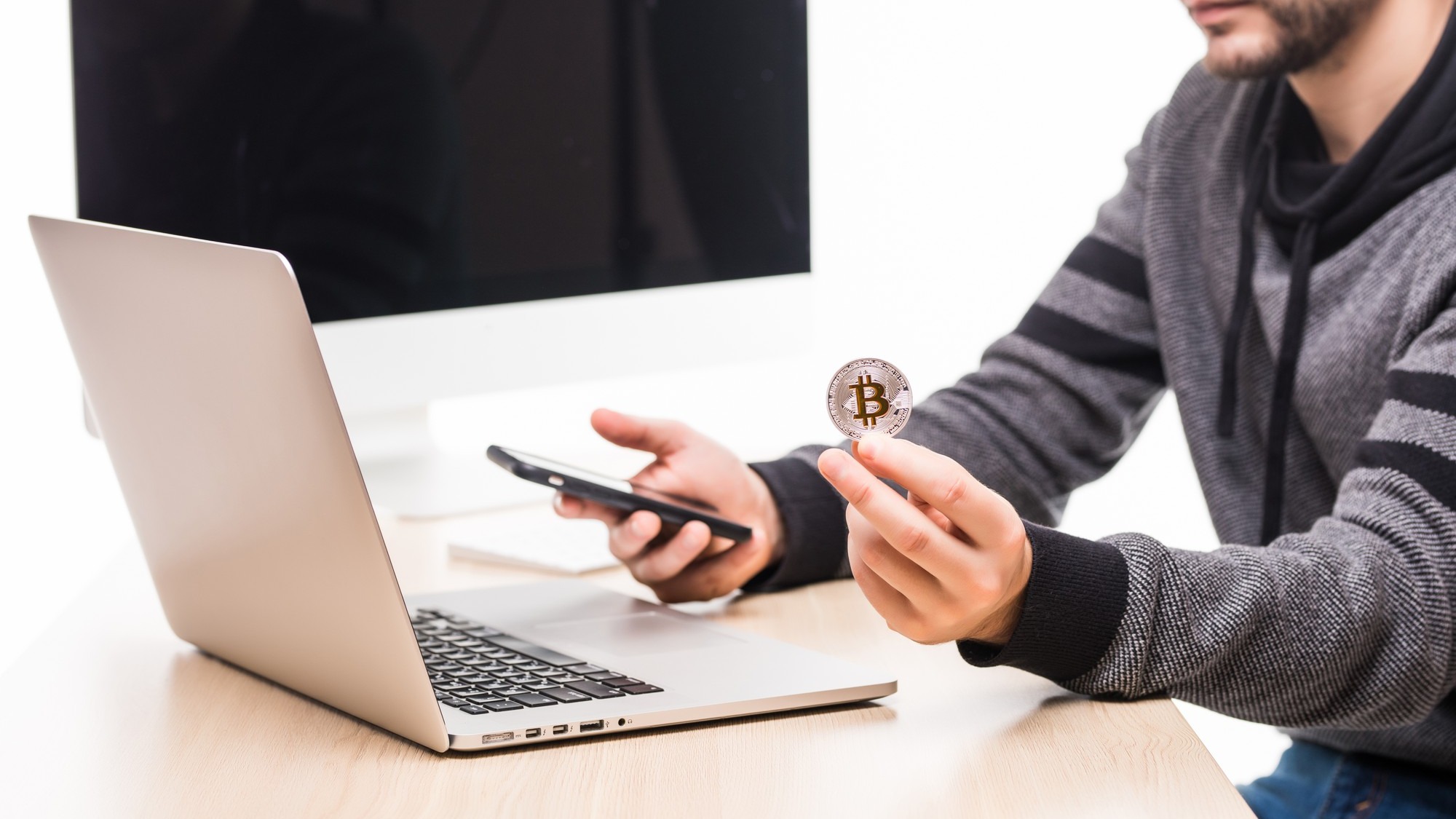 Close up of hands of man at working place woth laptop and monitor screen, holding phone and bitcoin