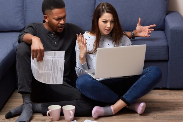 Indoor shot of multicultural couple spending free time at home and checking bills, dressed casually, sitting in front of laptop