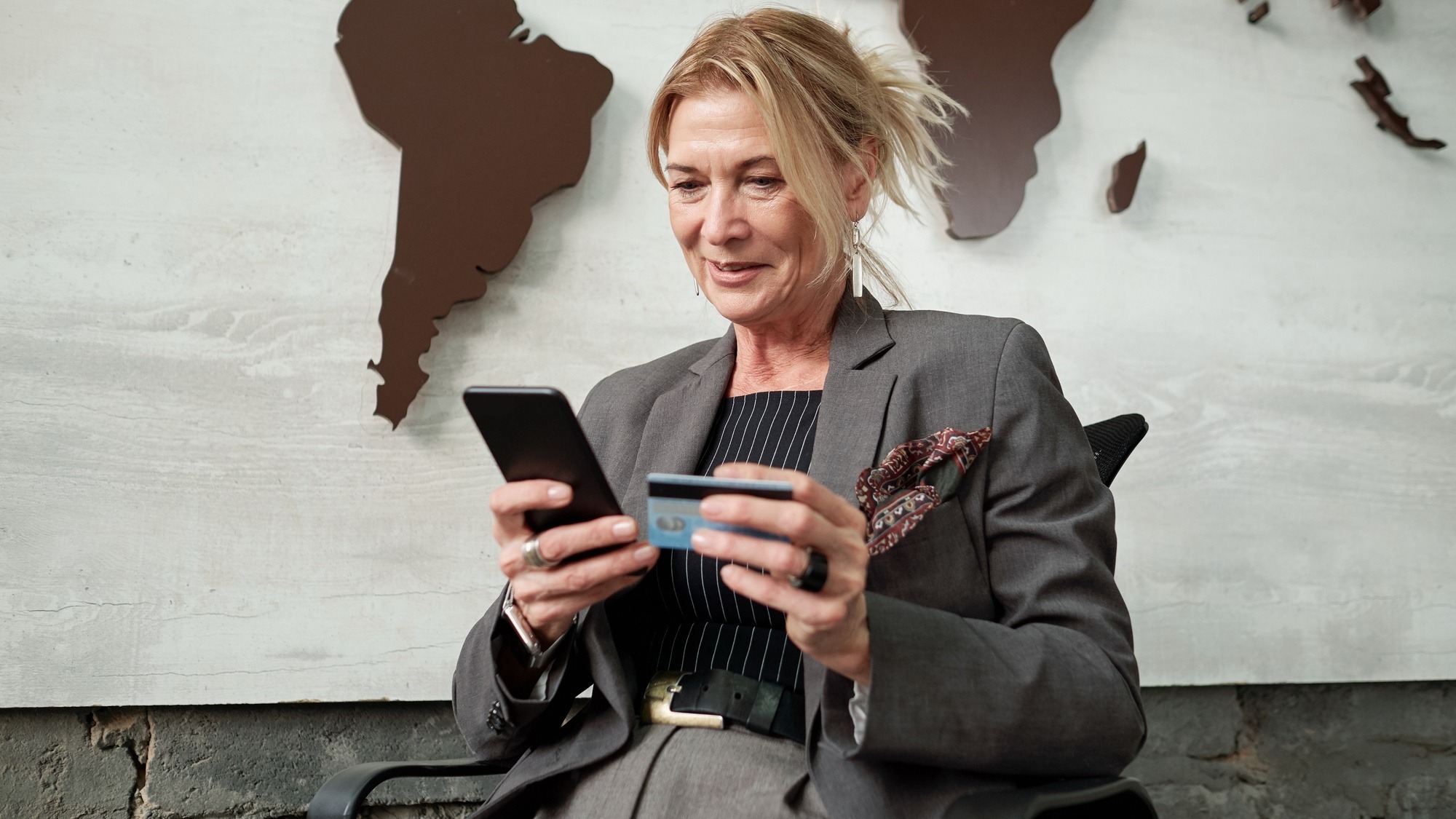 Content mature businesswoman sitting in chair and using smartphone and credit card while transferring money online