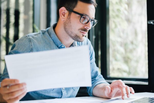 Serious pensive thoughtful focused young casual business accountant bookkeeper in office looking at and working with laptop and income tax return papers and documents