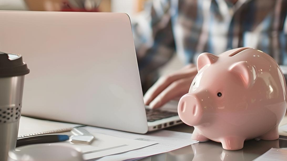 Piggy bank in soft focus on desk with blurred elderly man using laptop to fill out Internet pension application do accounting and paperwork at home manage personal finance or pay bills online