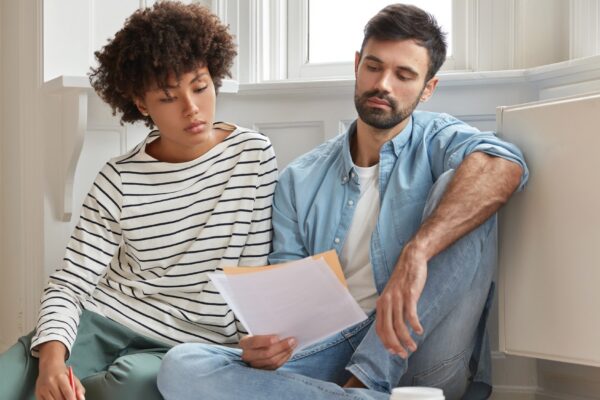 Male bearded accountant and his secretary work together in modern apartment, pose on wooden floor and discuss financial report, have serious gaze in papers, study analytics, feel comfort at home