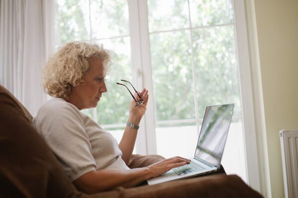 Elderly woman sitting in an armchair and holding her glasses while typing on a laptop.