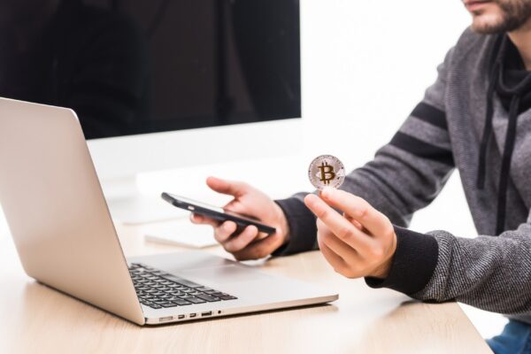 Close up of hands of man at working place woth laptop and monitor screen, holding phone and bitcoin
