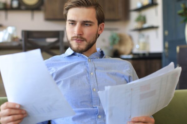 Concentrated young freelancer businessman sitting on sofa with laptop and examining documents.