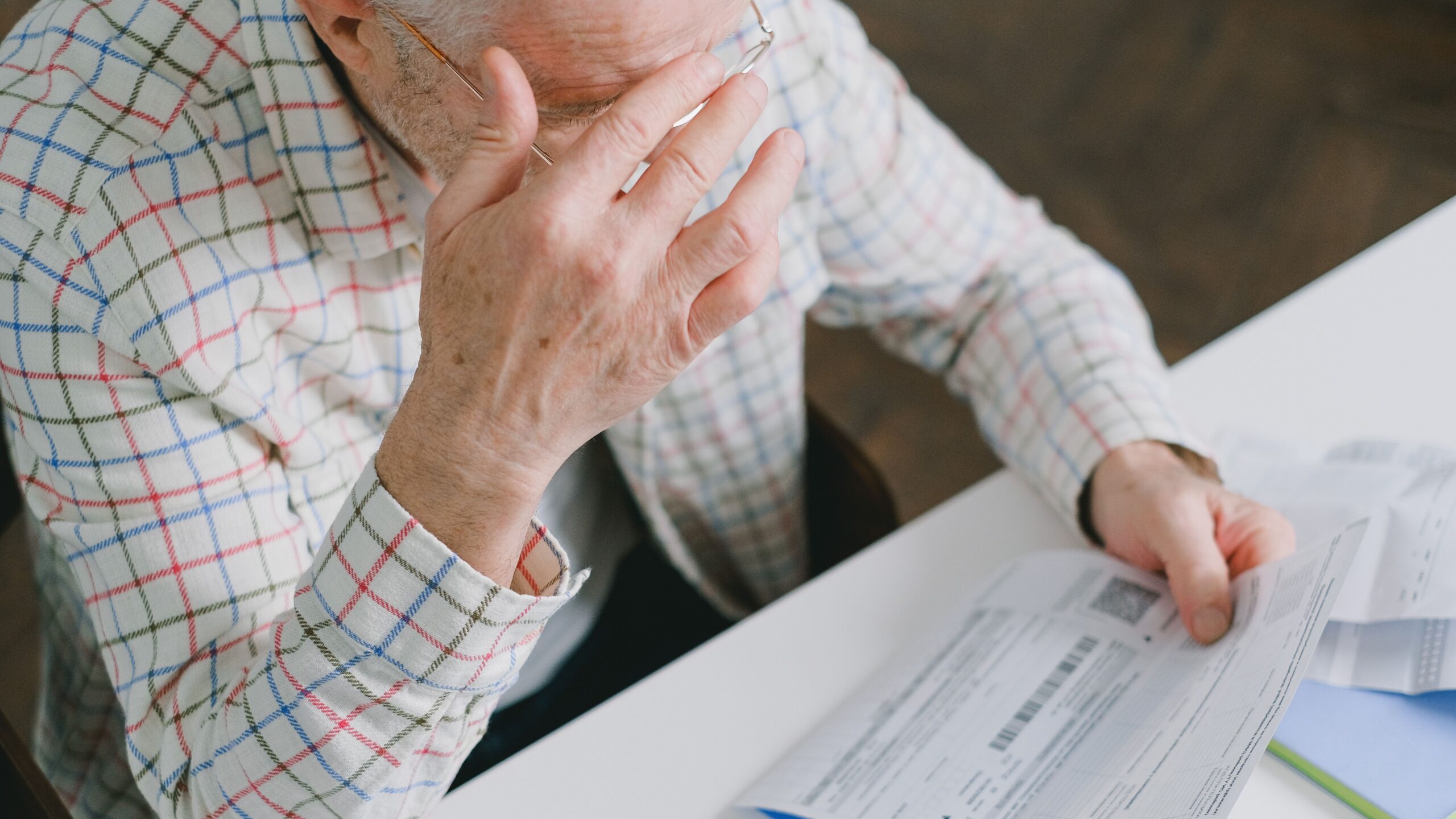 Elderly man in patterned shirt reading and holding bills at a home table, appearing focused.